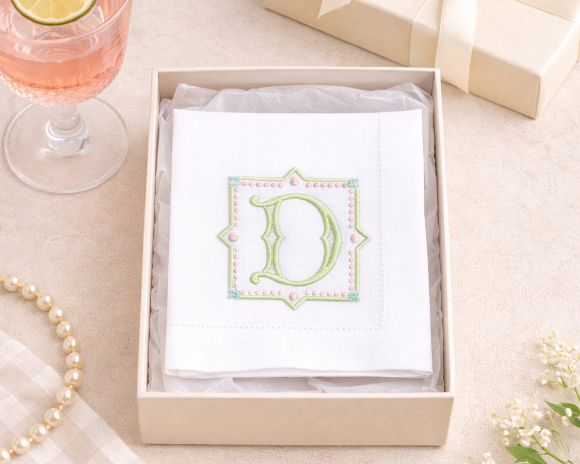 Boxed set with a card featuring a decorative letter 'D' on a table with a glass of pink drink and pearl necklace.