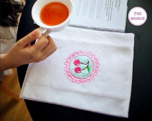 Hand holding a cup of tea over a white handkerchief with an embroidered design, branded 'The Hankie'.