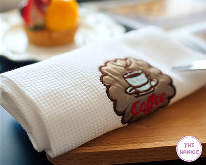 White towel with embroidered design of a coffee cup and 'Coffee' text on a wooden surface.