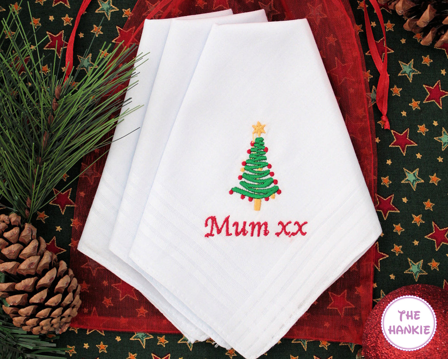 White hankies with embroidered Christmas tree and 'Mum xx' text on a festive background with pine cones and stars.