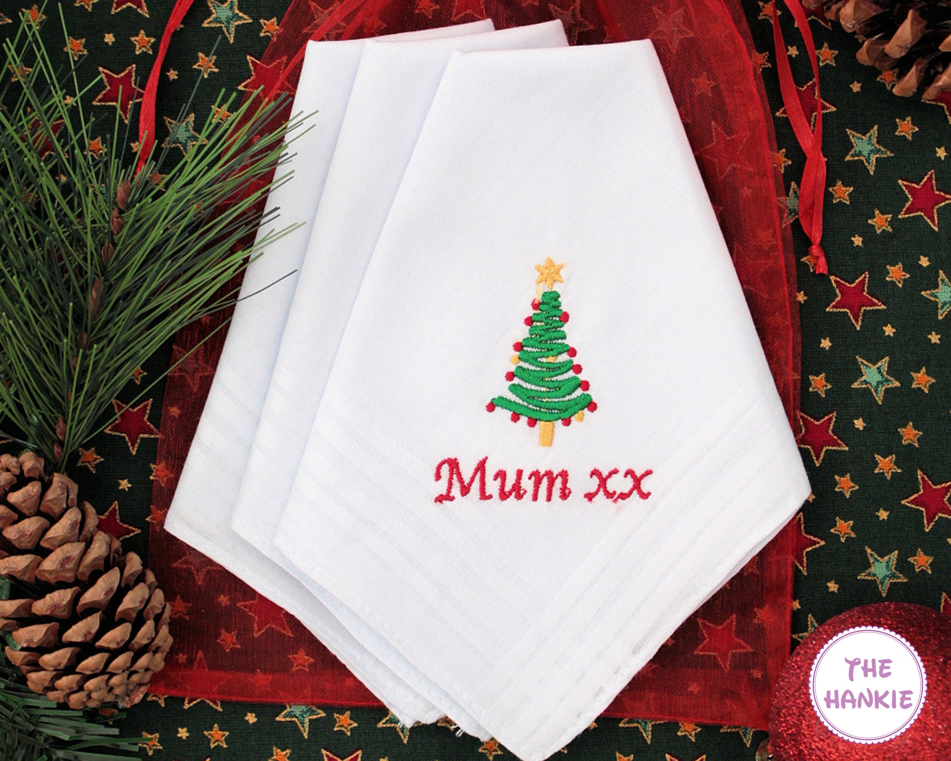 White hankies with embroidered Christmas tree and 'Mum xx' text on a festive background with pine cones and stars.