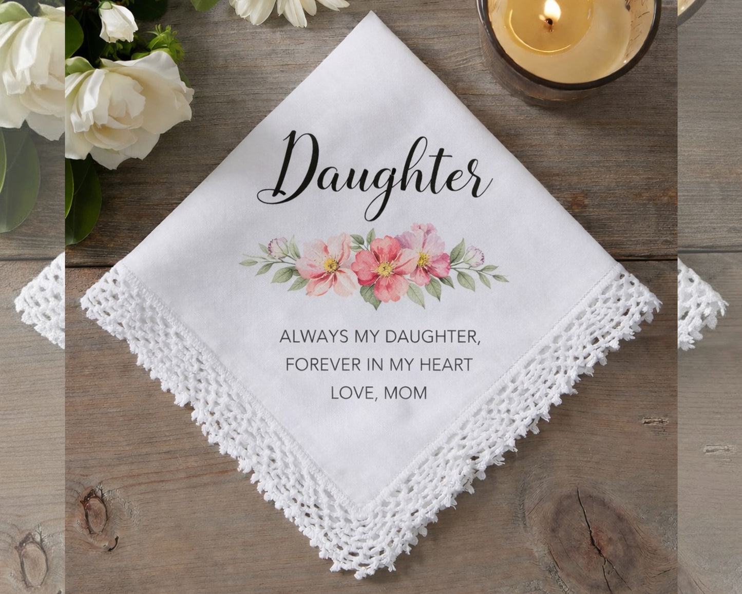 White handkerchief with floral design and text on a wooden surface with flowers and a candle.