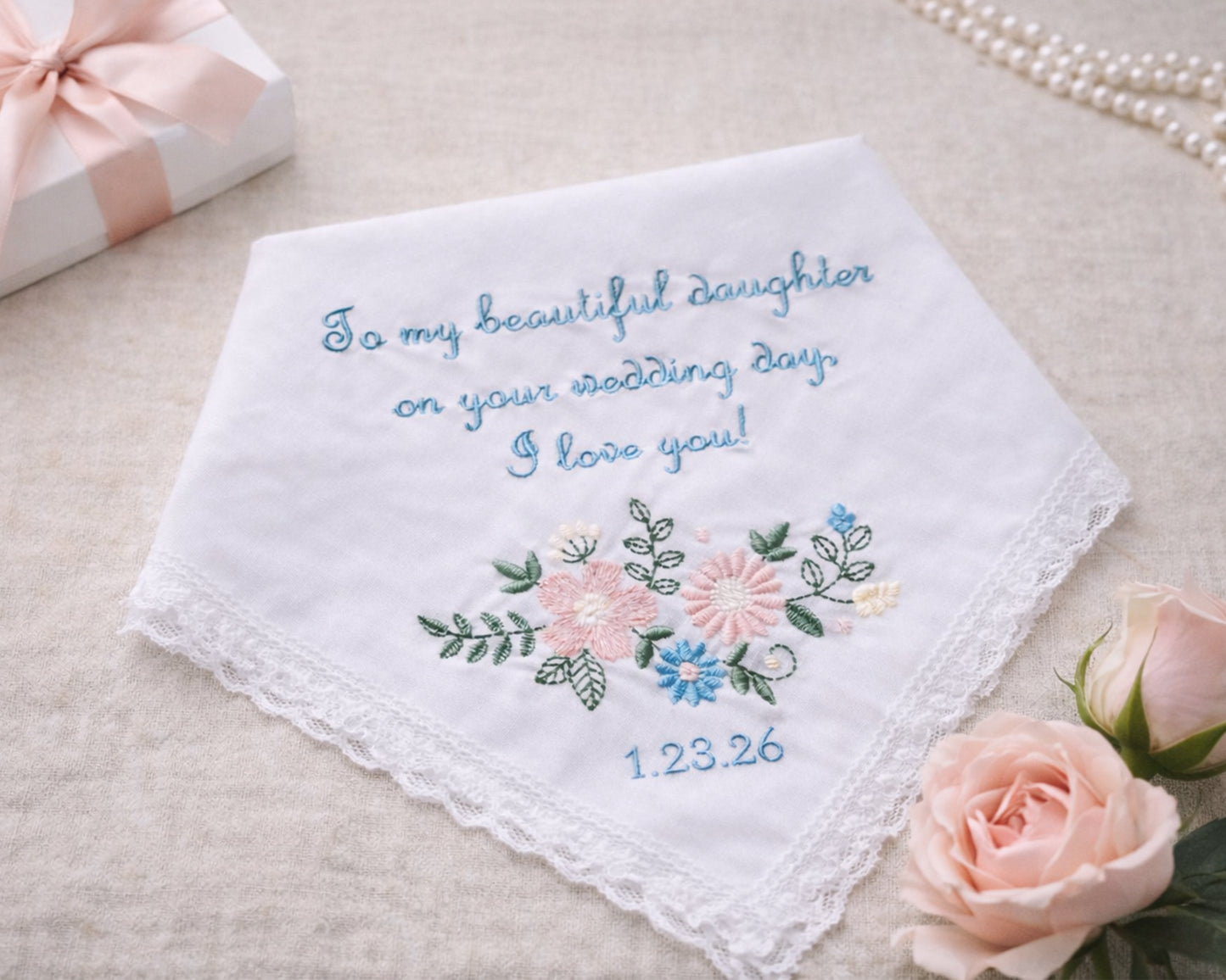 Embroidered wedding handkerchief with floral design and text on a beige surface with roses and a gift box.