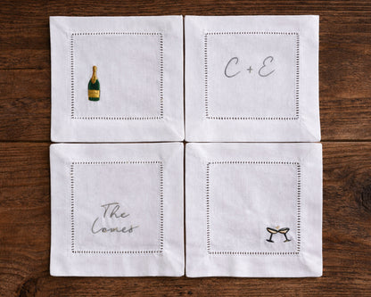 Four white cocktail napkins with embroidered designs on a wooden surface