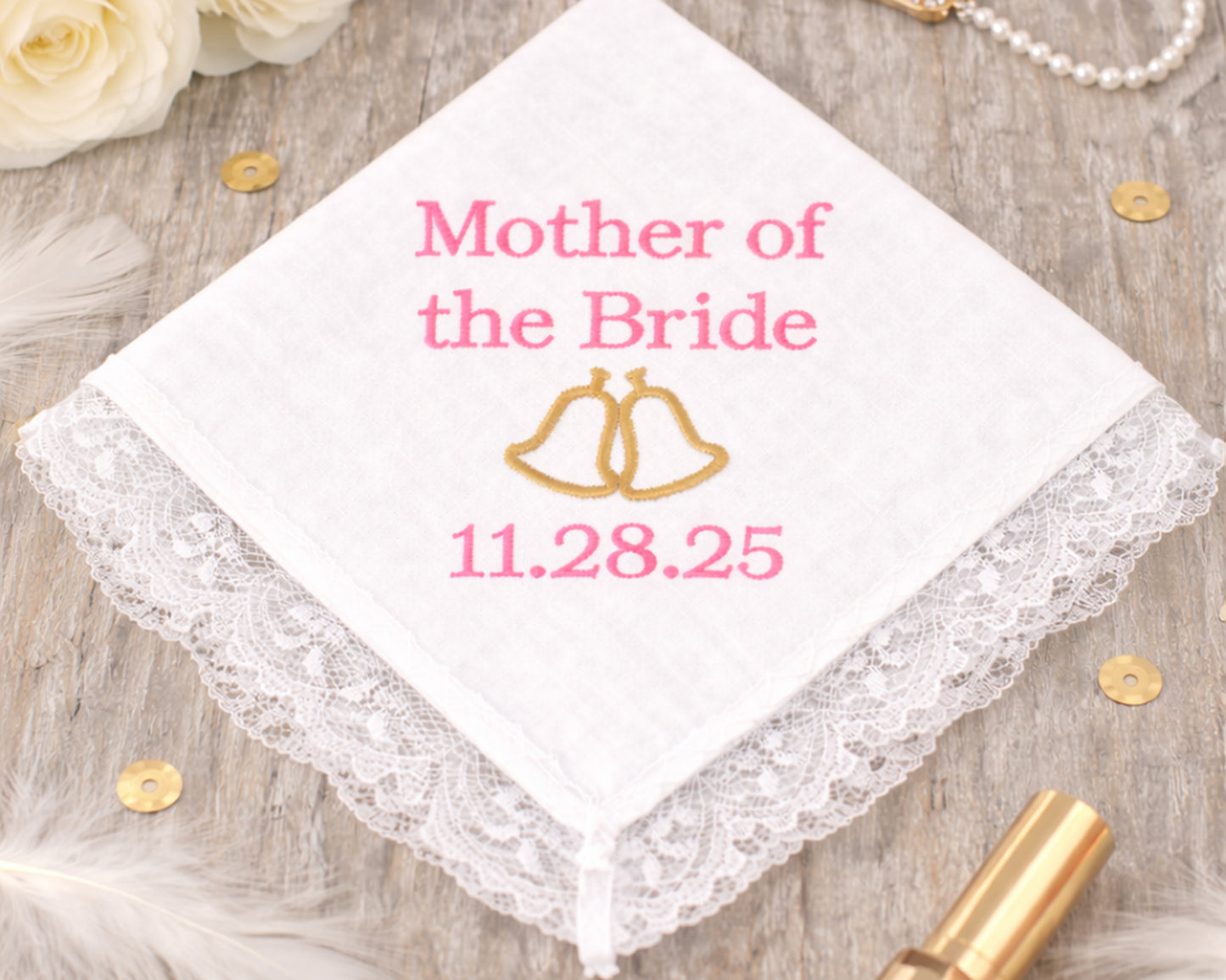 Handkerchief with 'Mother of the Bride' and wedding date on a wooden surface with flowers and jewelry.
