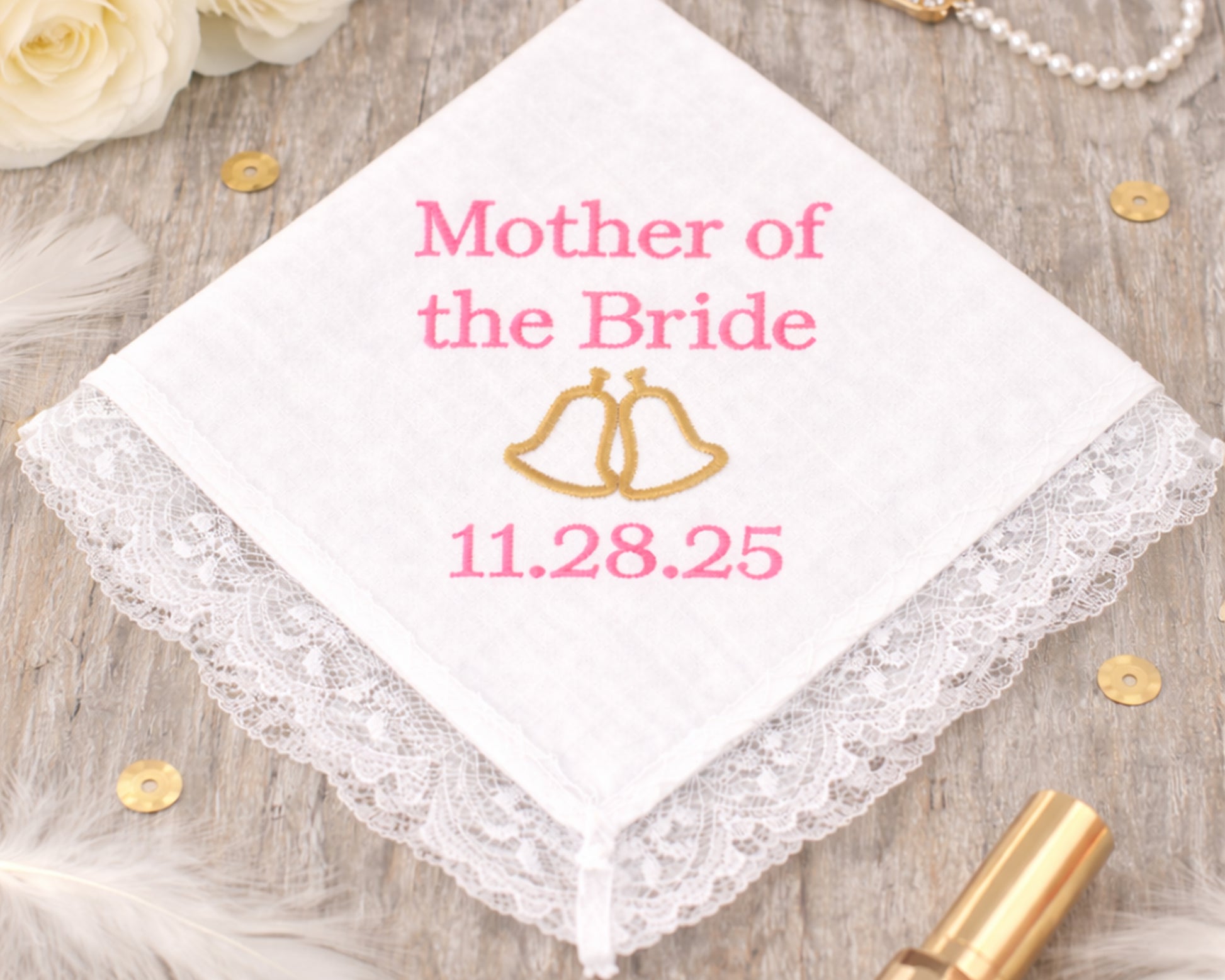 Handkerchief with 'Mother of the Bride' and wedding date on a wooden surface with flowers and jewelry.