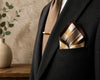 Man wearing a black suit with a patterned pocket square and brown tie, standing against a neutral wall.