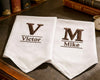 Two white napkins with embroidered initials and names on a wooden surface.