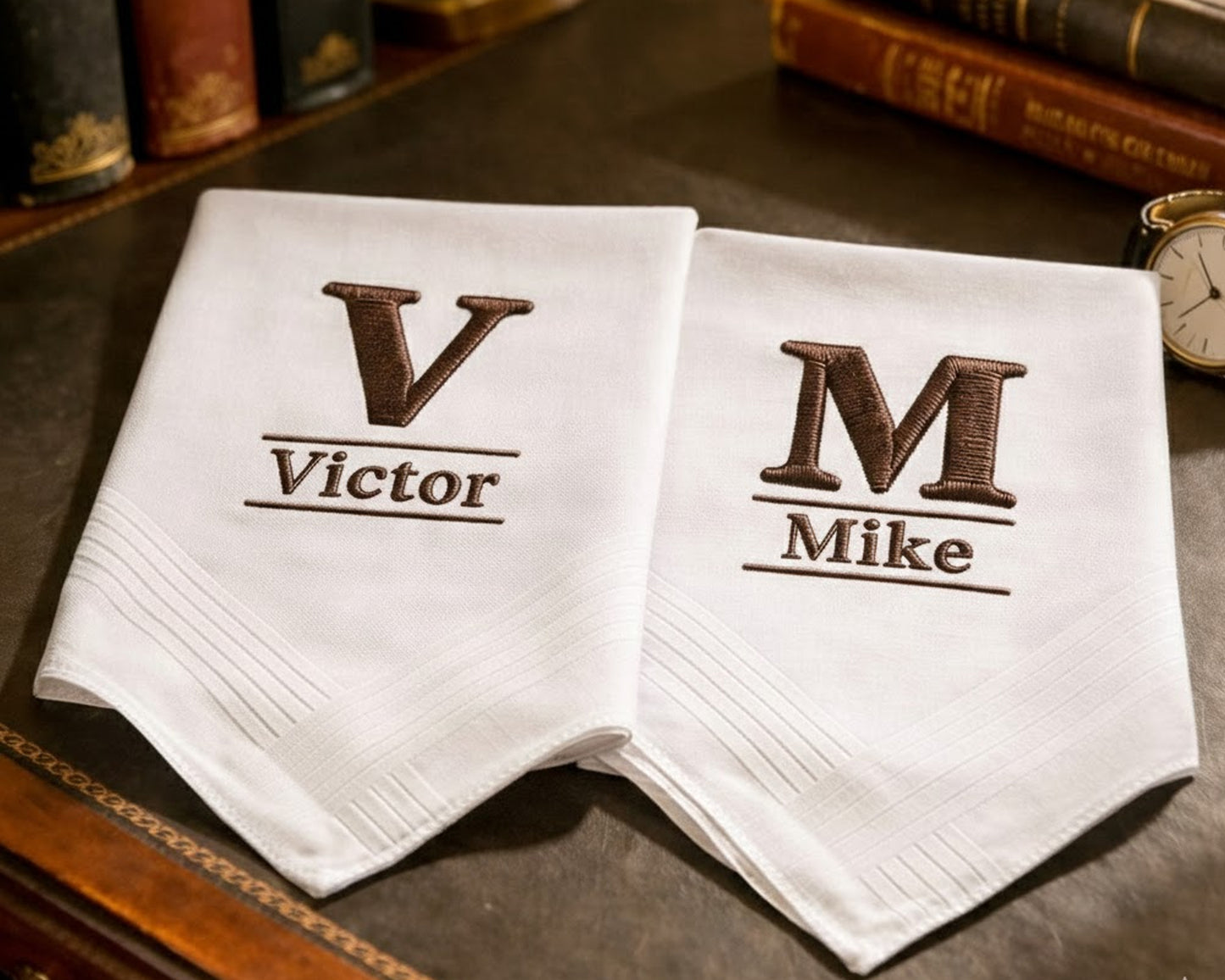 Two white napkins with embroidered initials and names on a wooden surface.