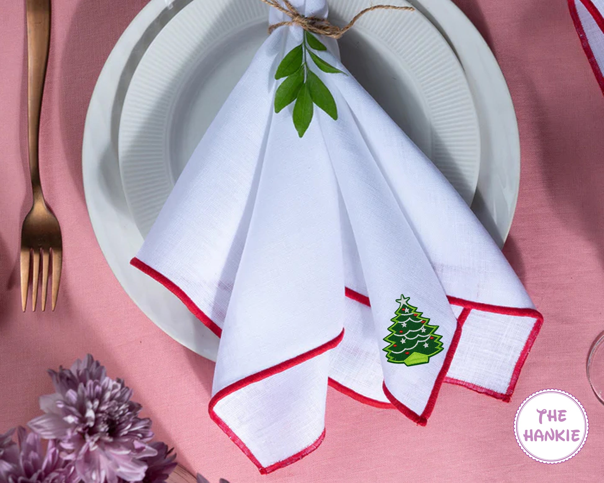 Napkin with green tree design on a pink tablecloth with 'The Hankie' branding.