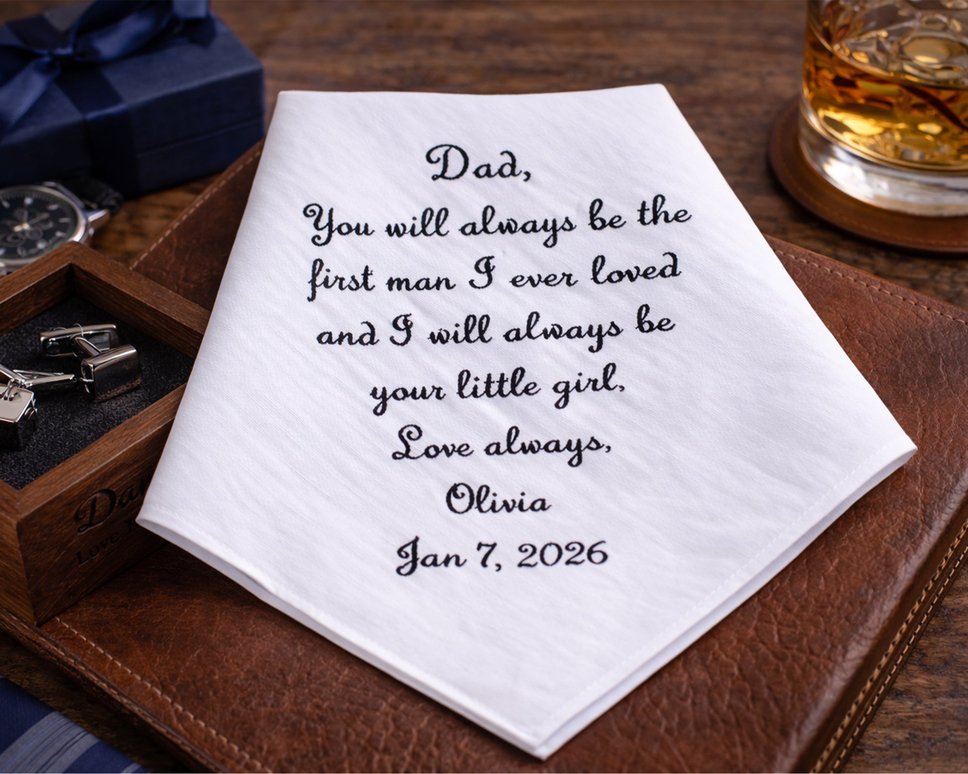 Personalized handkerchief with a heartfelt message on a wooden surface with a glass of whiskey and jewelry box.