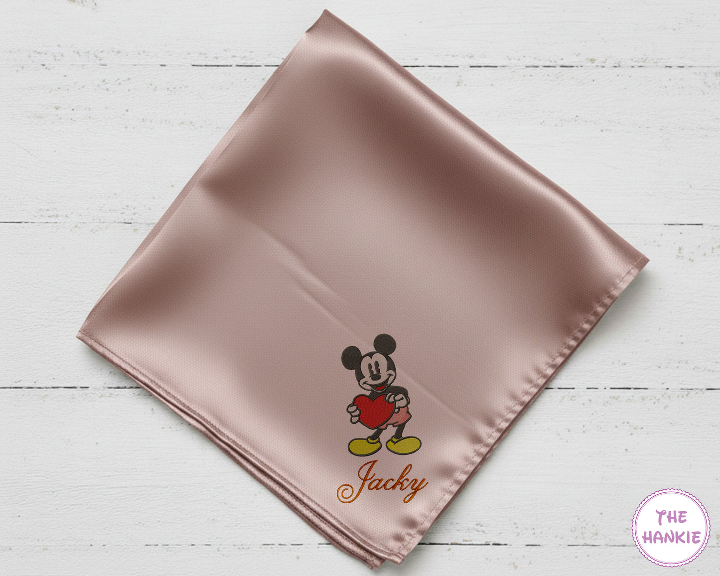 Pink satin square with embroidered character and 'Jacky' text on a light wooden surface, branded 'The Hankie'.