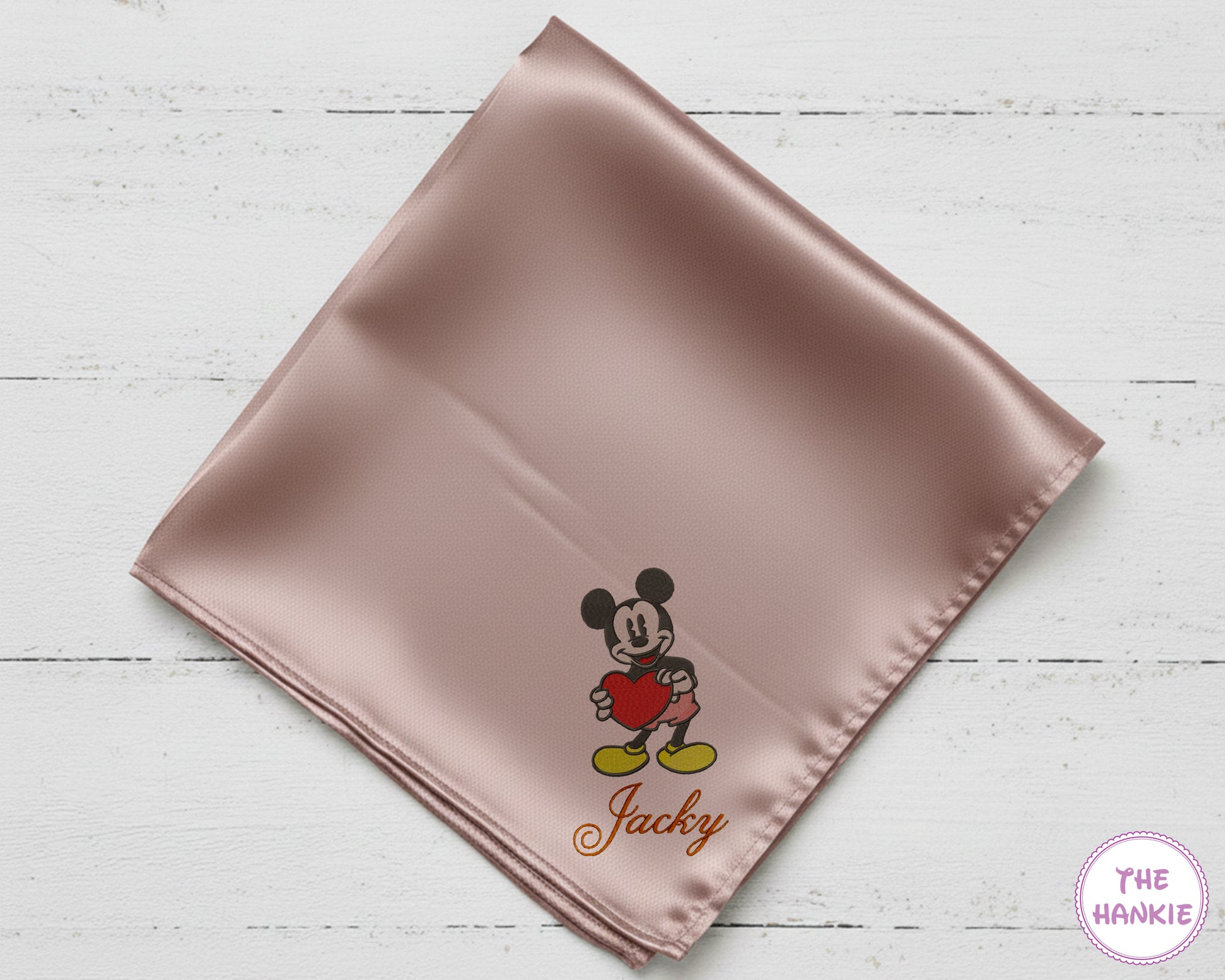 Pink satin square with embroidered character and 'Jacky' text on a light wooden surface, branded 'The Hankie'.