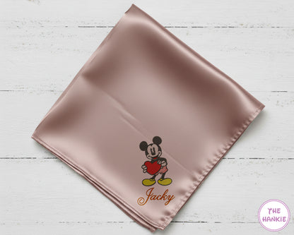 Pink satin square with embroidered character and 'Jacky' text on a light wooden surface, branded 'The Hankie'.