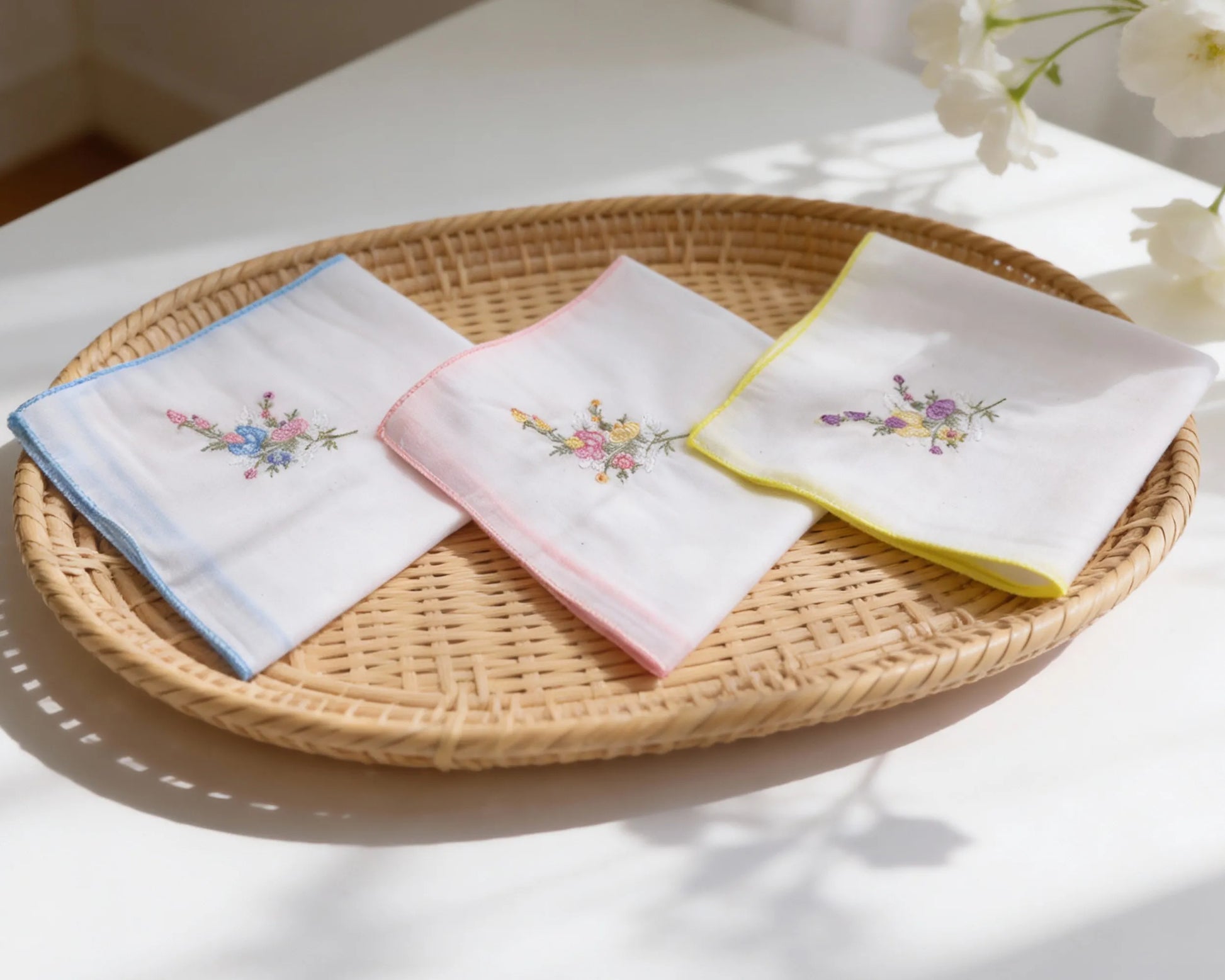 Three embroidered handkerchiefs on a woven tray with flowers in the background