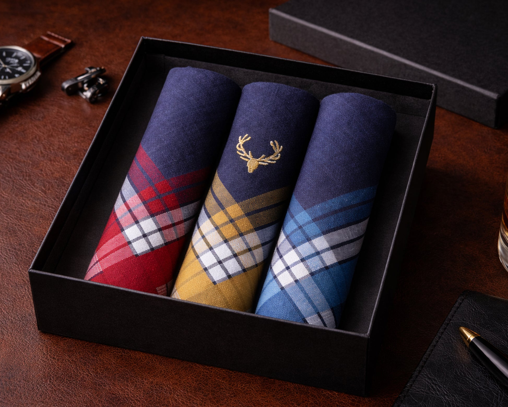 Three plaid pocket squares in a black box on a wooden surface with a watch and pen.