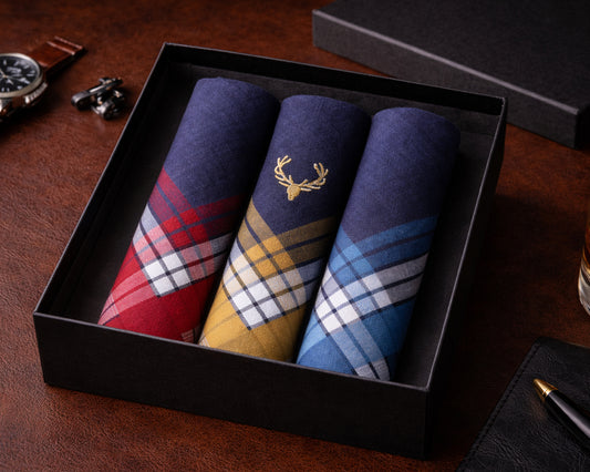 Three plaid pocket squares in a black box on a wooden surface with a watch and pen.