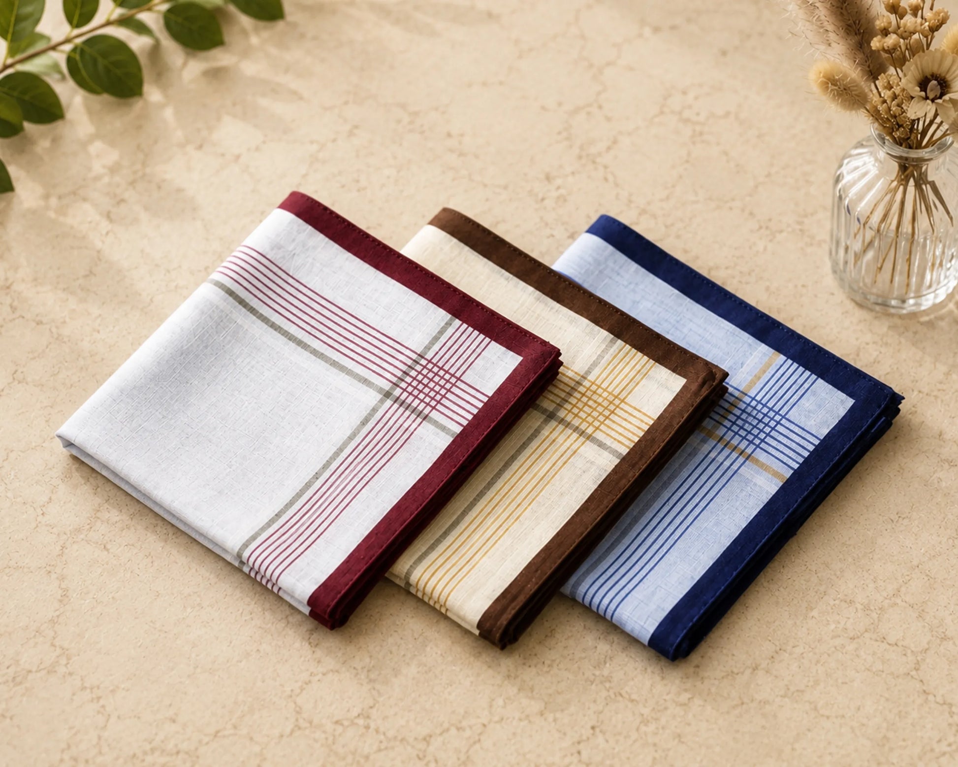 Three plaid pocket squares in different colors on a beige surface with a vase and leaves in the background.