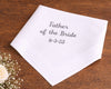 White handkerchief with 'Father of the Bride' and date on a wooden surface with flowers.