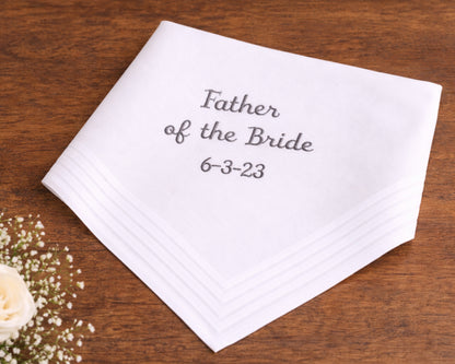 White handkerchief with 'Father of the Bride' and date on a wooden surface with flowers.