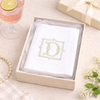 White handkerchief with a decorative monogram in a box, accompanied by a glass of pink drink and gift box.