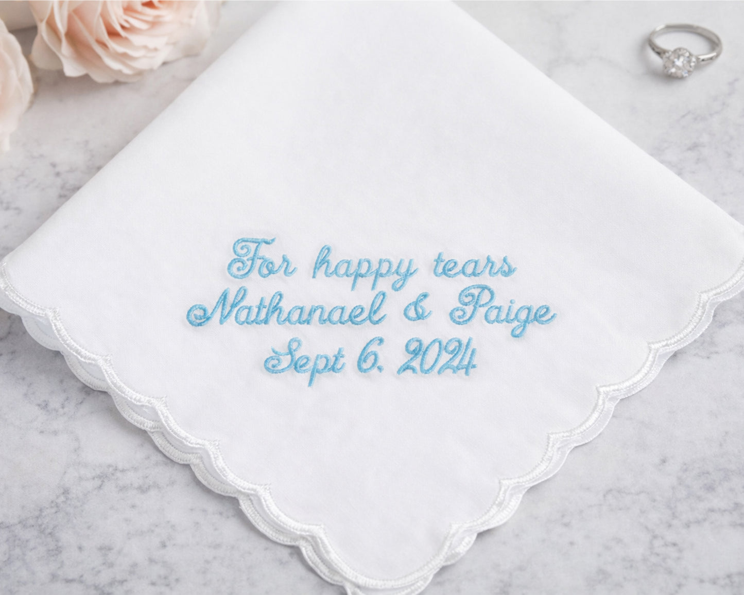 White handkerchief with blue embroidered text on a marble surface with flowers and a ring.