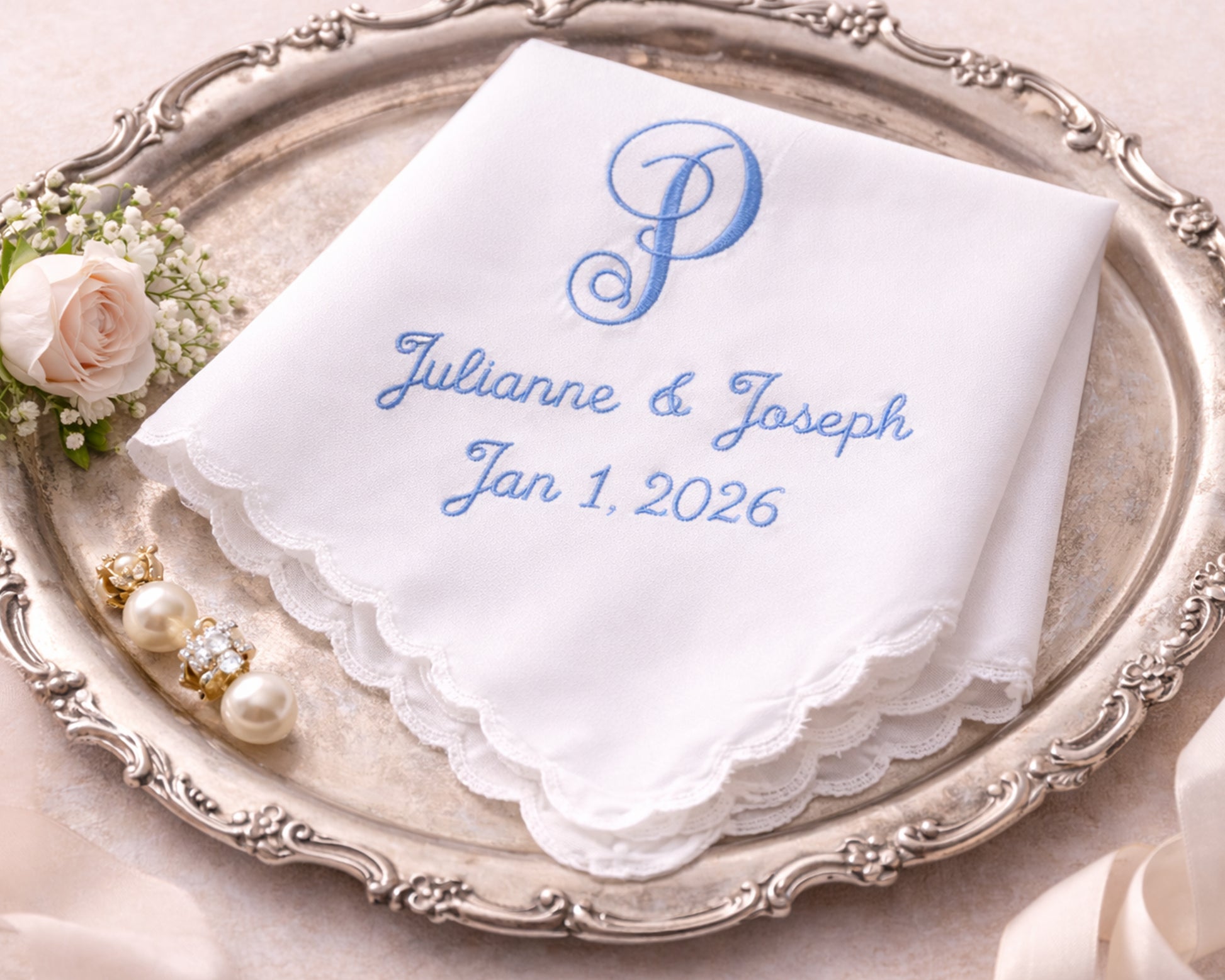 White handkerchief with blue embroidery on a decorative silver tray with flowers and jewelry.