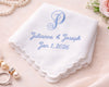 White handkerchief with blue embroidery of names and date, surrounded by jewelry and flowers on a light background