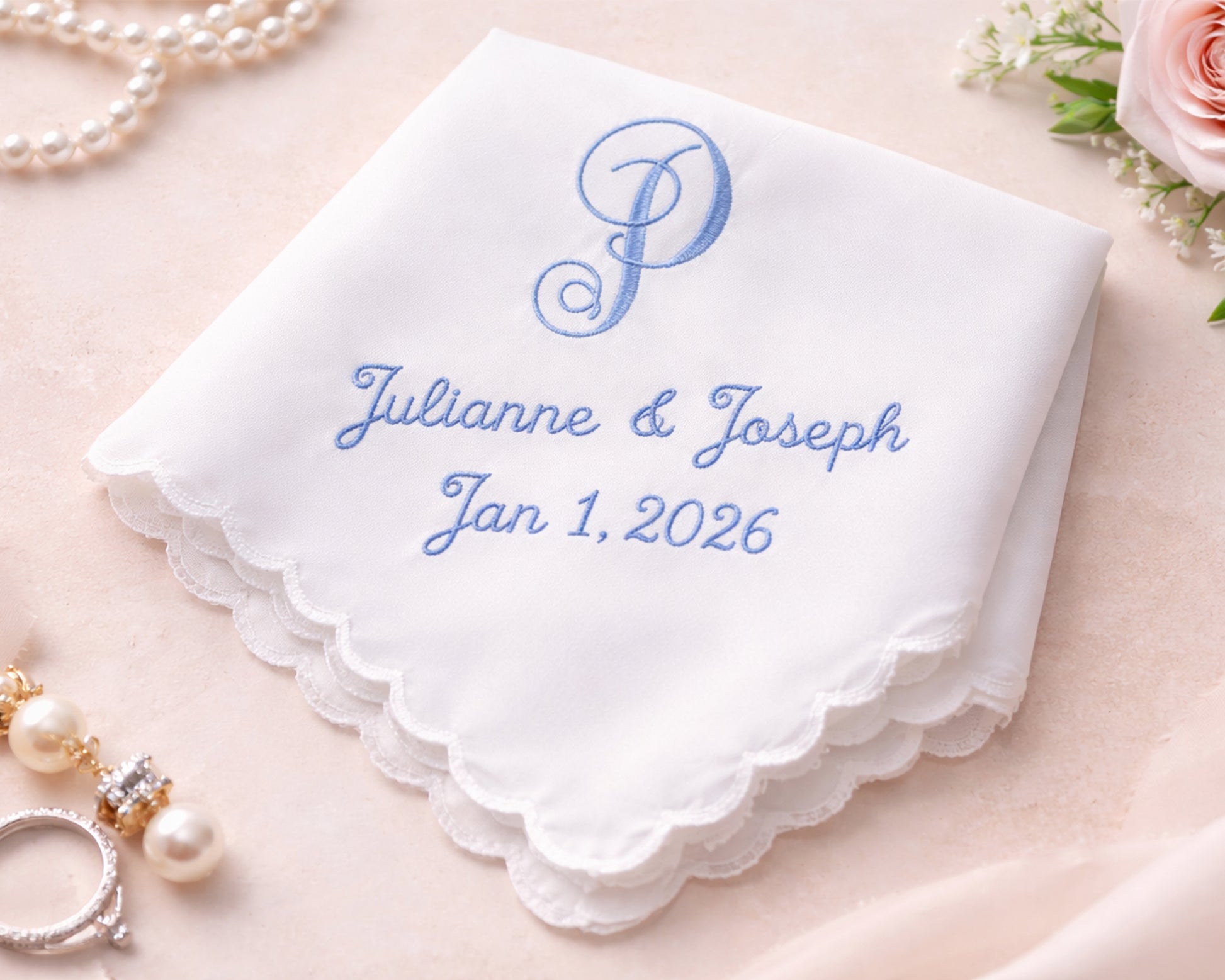 White handkerchief with blue embroidery of names and date, surrounded by jewelry and flowers on a light background