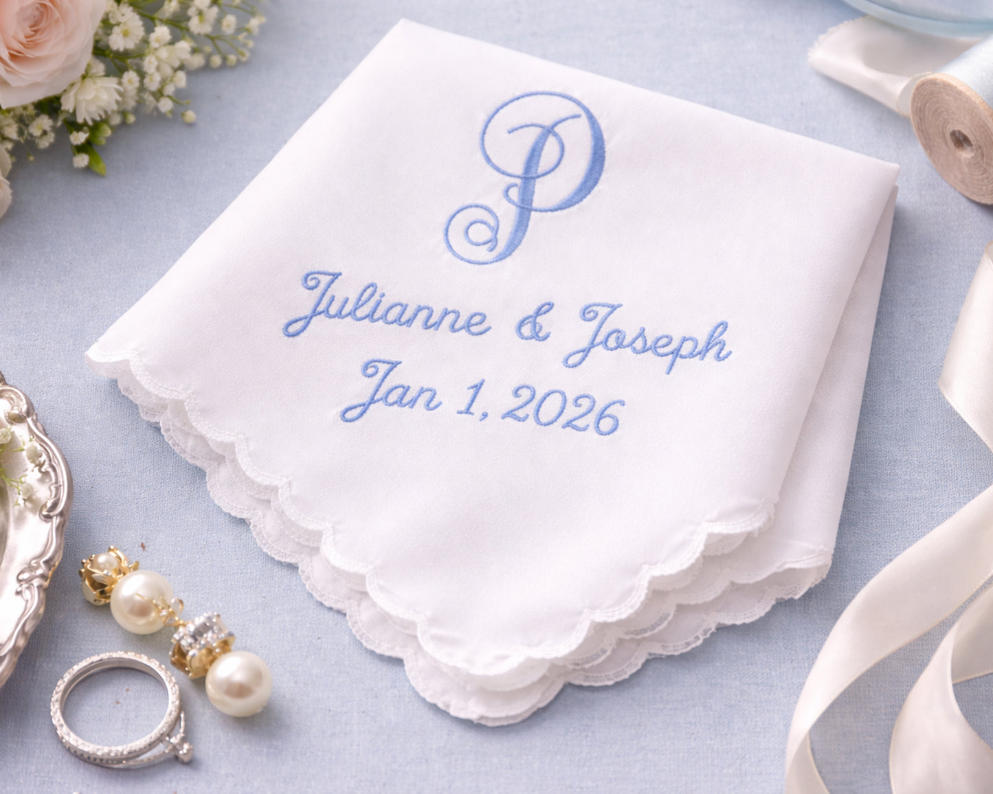 White handkerchief with blue embroidery of 'Julianne & Joseph Jan 1, 2026' on a light blue background with wedding accessories.