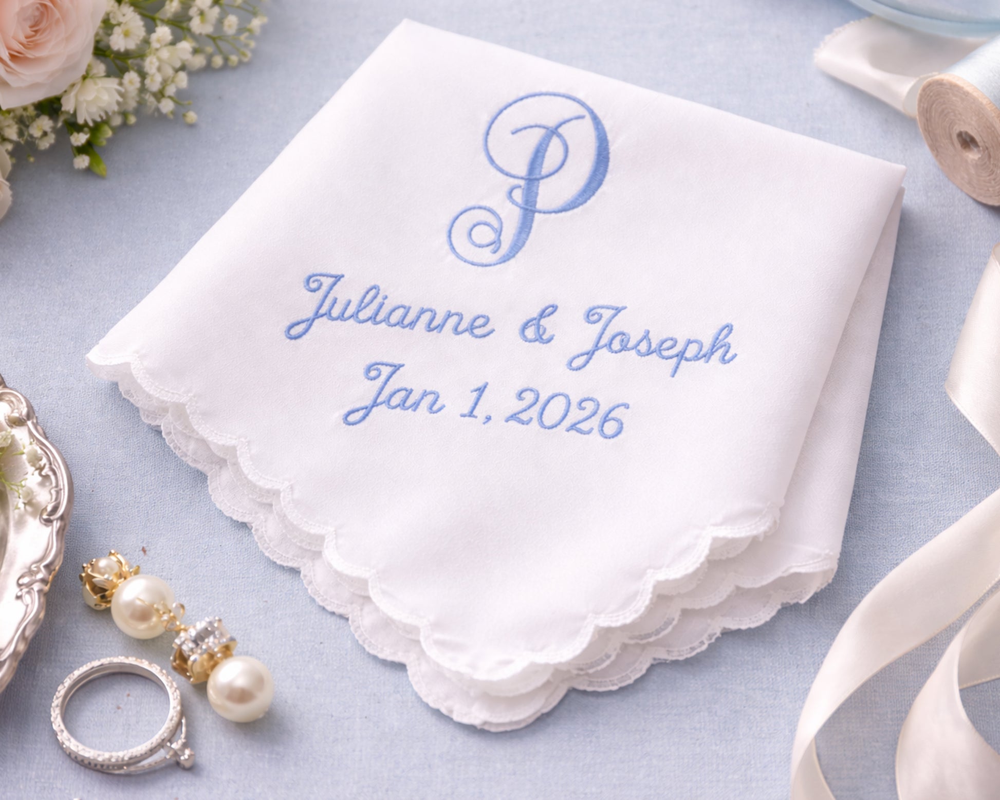 White handkerchief with blue embroidery of 'Julianne & Joseph Jan 1, 2026' on a light blue background with wedding accessories.