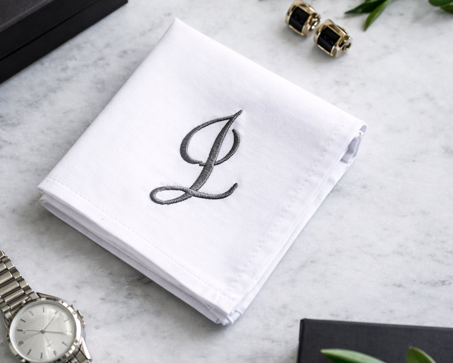White handkerchief with embroidered letter 'L' on a marble surface with jewelry and a watch.