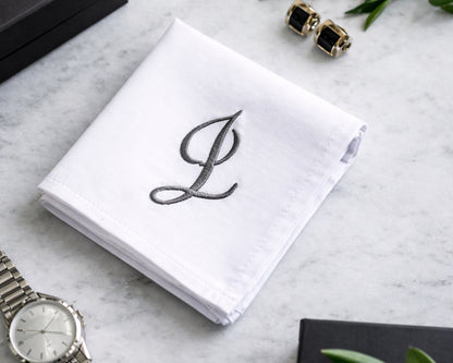 White handkerchief with embroidered letter 'L' on a marble surface with jewelry and a watch.