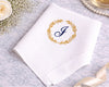 White handkerchief with gold laurel wreath and blue 'J' monogram on a marble surface with decorative elements.