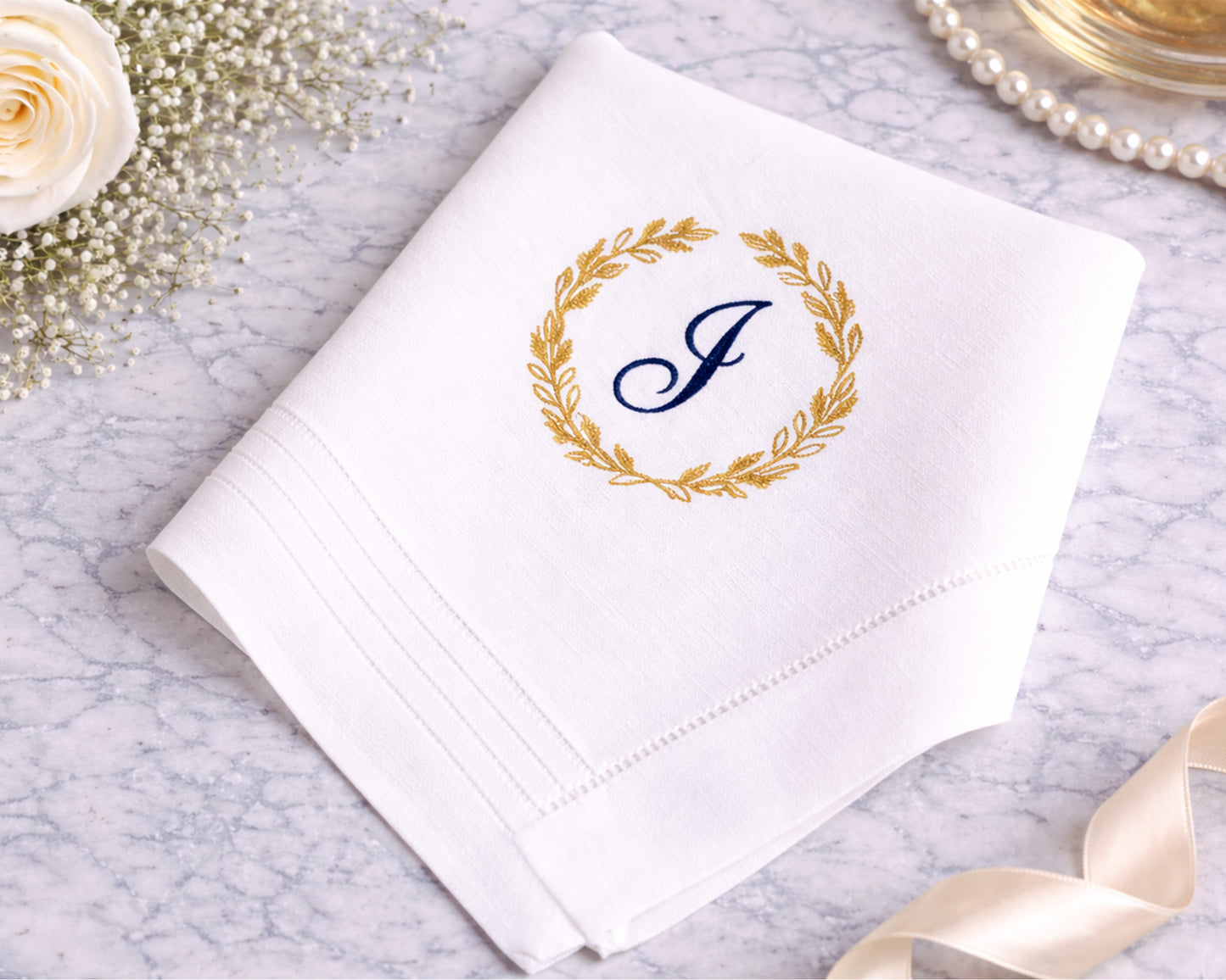 White handkerchief with gold laurel wreath and blue 'J' monogram on a marble surface with decorative elements.