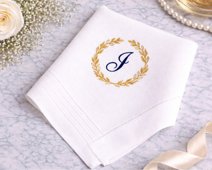 White handkerchief with gold laurel wreath and blue 'J' monogram on a marble surface with decorative elements.