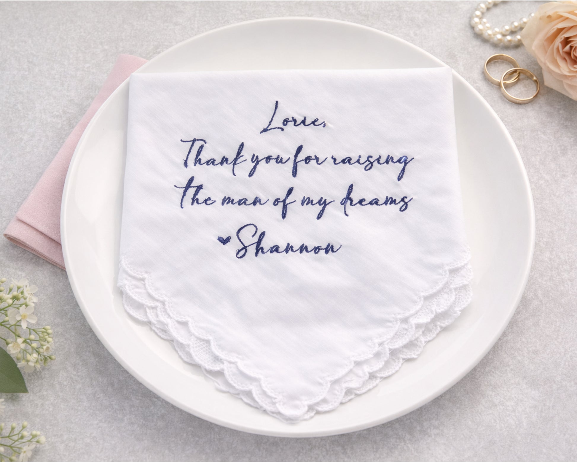 White handkerchief with personalized text on a white plate, surrounded by flowers and jewelry.