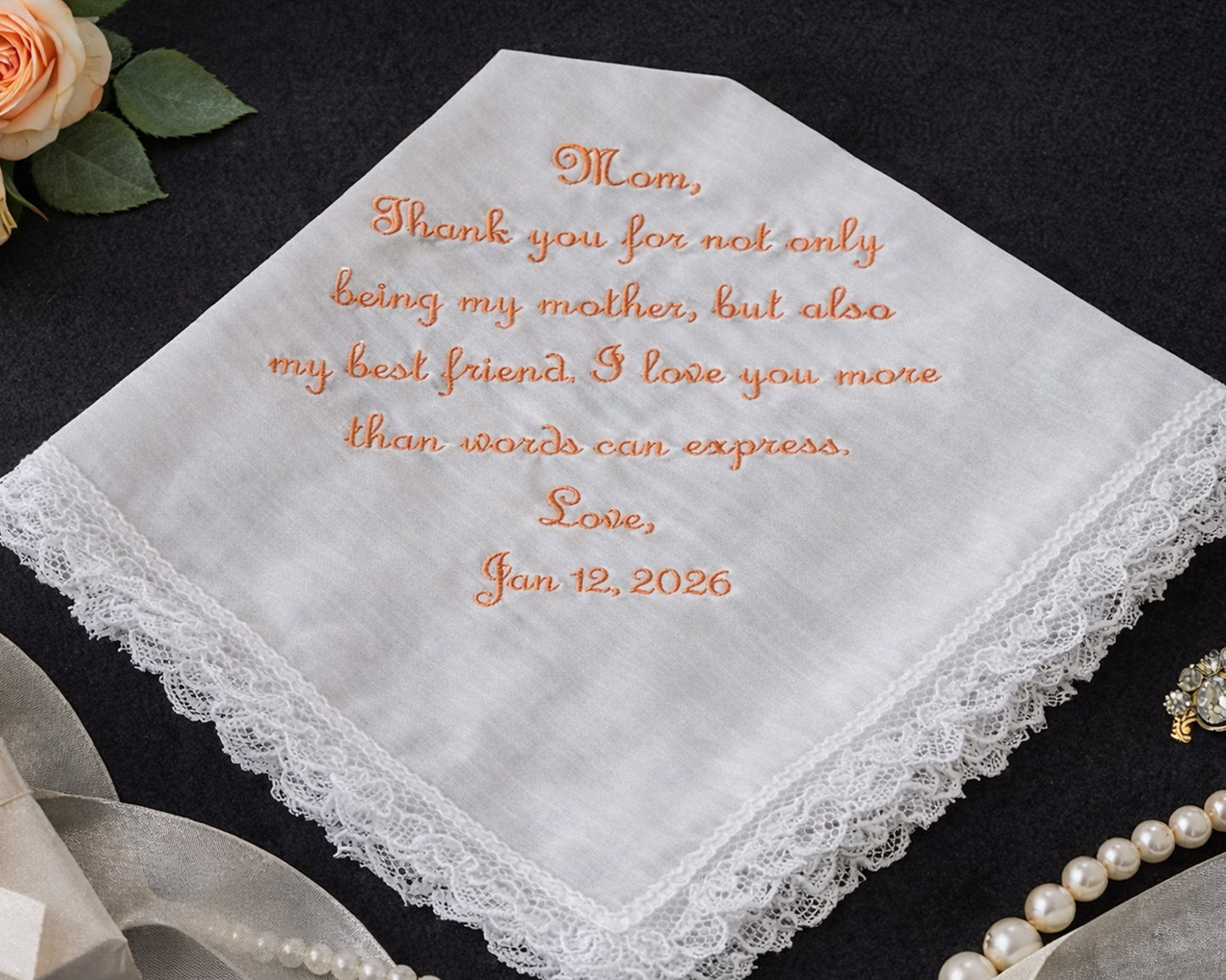 White handkerchief with embroidered message on a dark surface with flowers and jewelry.