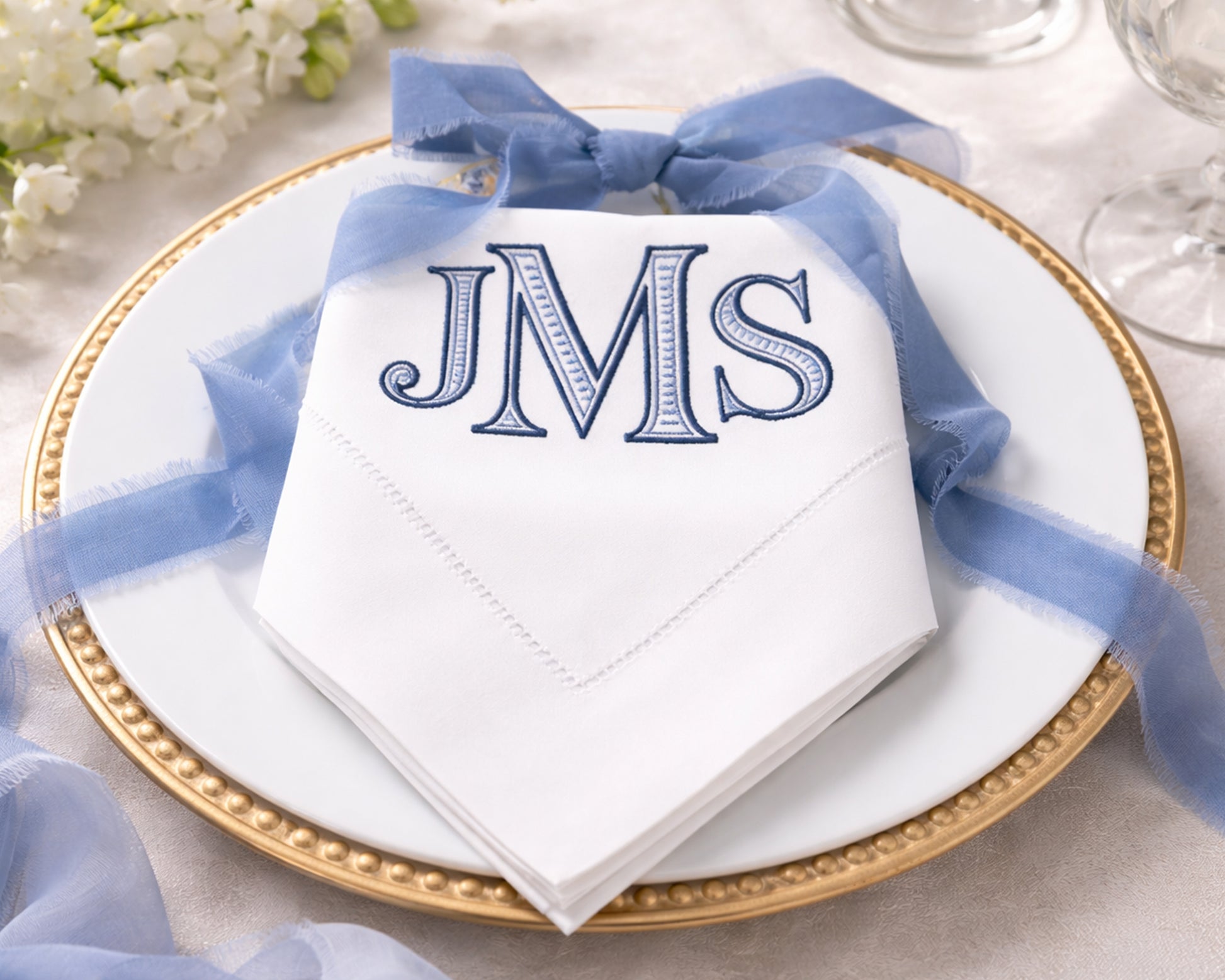 White napkin with 'JMS' monogram on a gold plate with blue ribbon