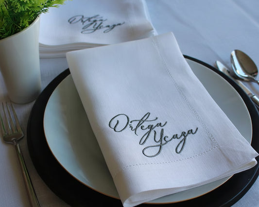White napkin with 'Ortega Ycaza' embroidery on a table setting with cutlery and plates.