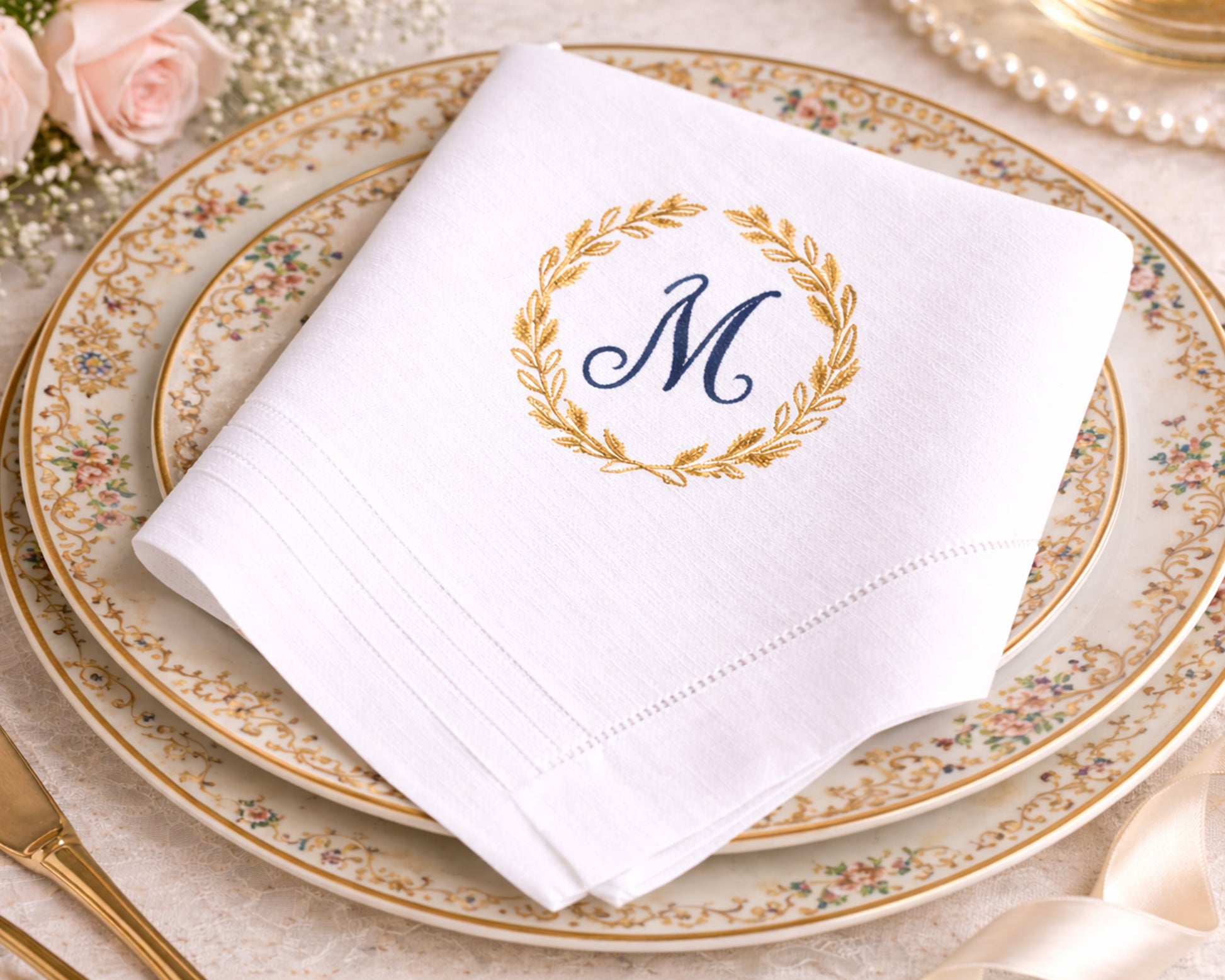 White napkin with gold wreath and blue 'M' monogram on a decorative plate.