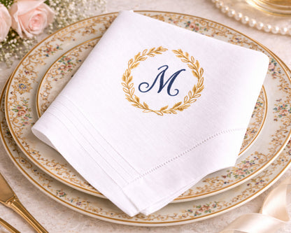 White napkin with gold wreath and blue 'M' monogram on a decorative plate.