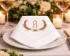 White napkin with gold monogram 'R' on a dinner setting with flowers and candles.