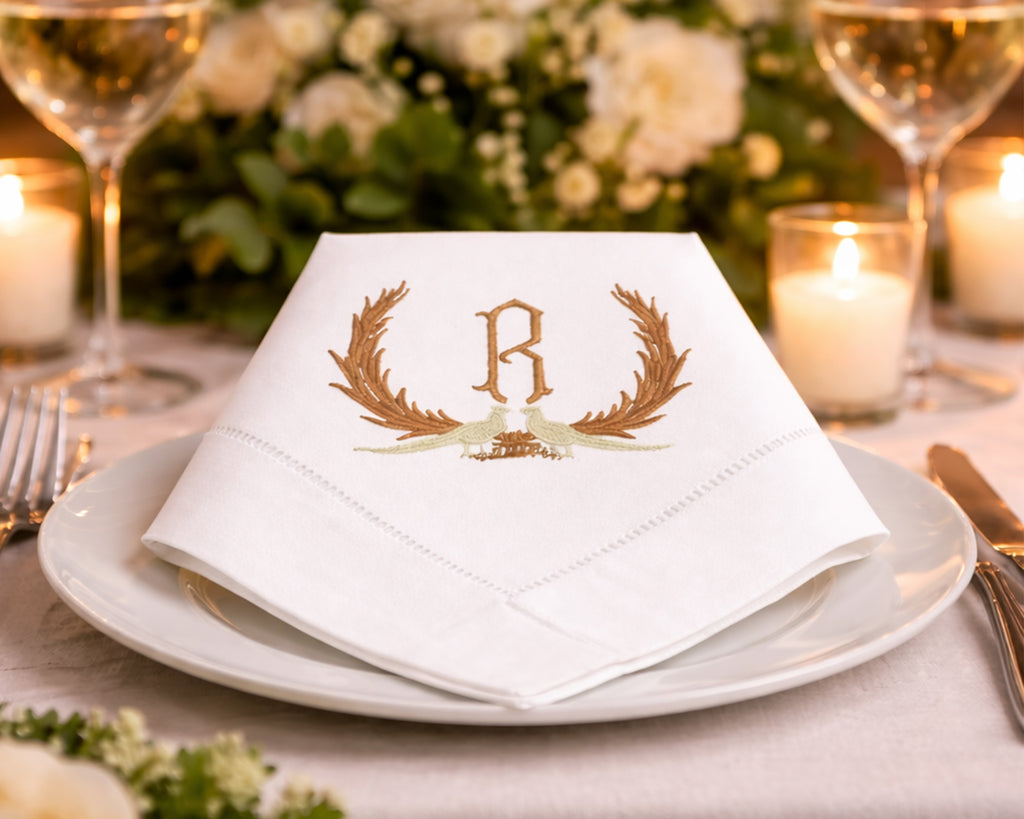 White napkin with gold monogram 'R' on a dinner setting with flowers and candles.