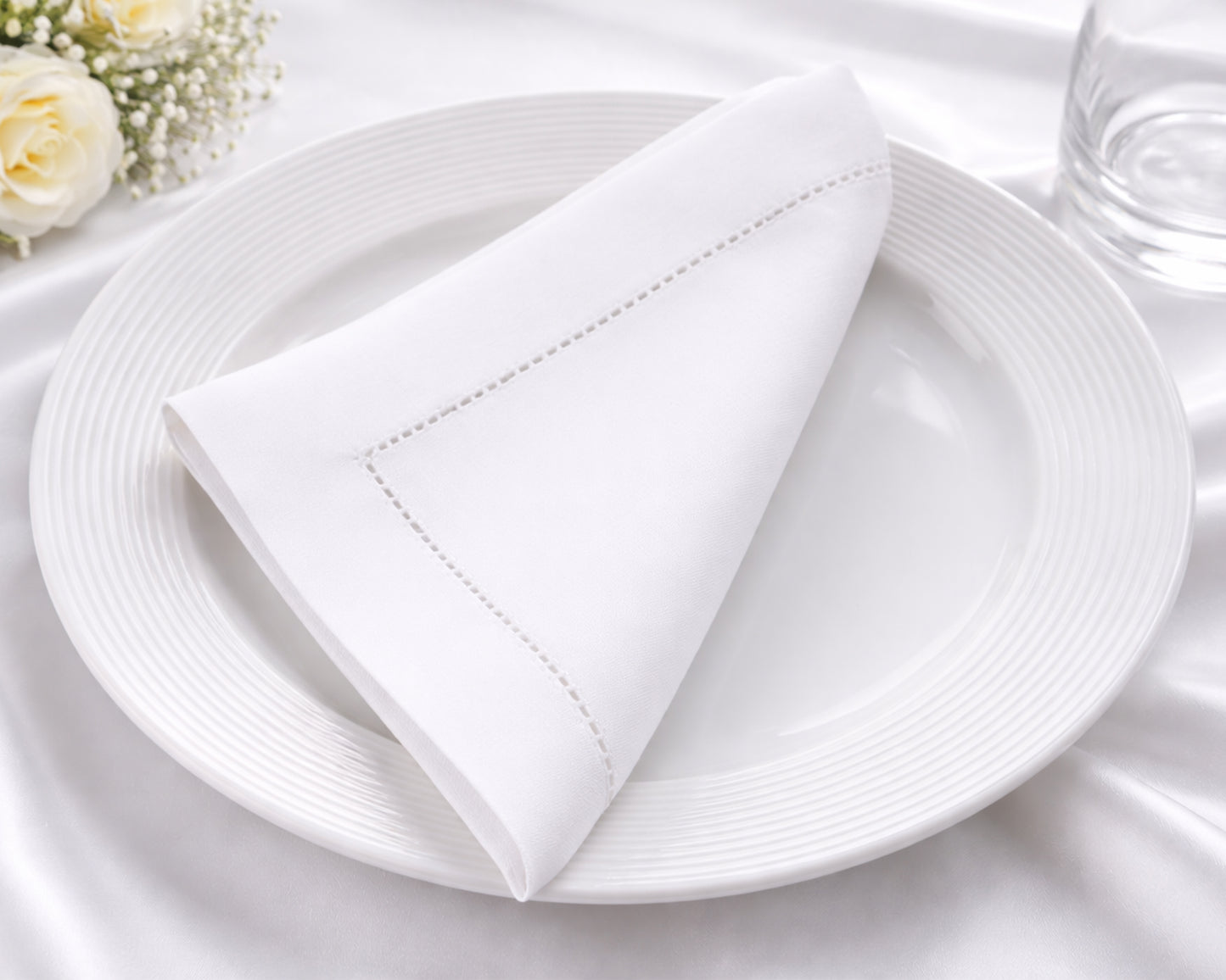 White plate with a folded white napkin on a white tablecloth, with flowers in the corner.