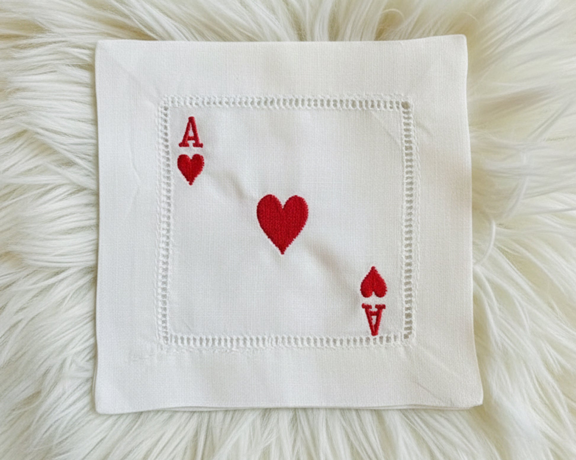 A single white cotton hemstitch napkin featuring a large center heart and "A" symbols for the Ace of Hearts in vibrant red machine embroidery.