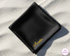 A luxury black satin handkerchief featuring the name "Amelia" custom-embroidered in a vibrant yellow script on a white sand beach.