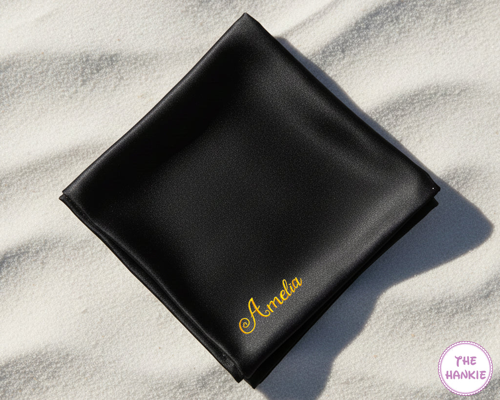 A luxury black satin handkerchief featuring the name "Amelia" custom-embroidered in a vibrant yellow script on a white sand beach.