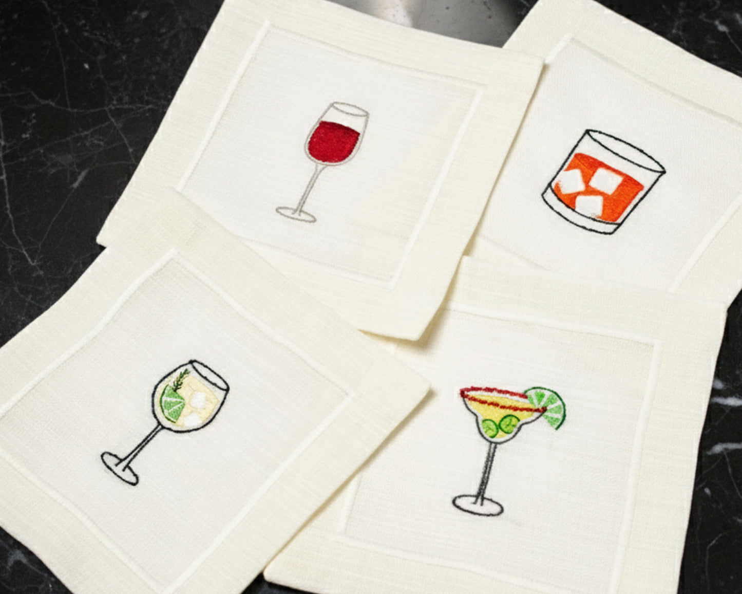 A variety of cream cotton napkins featuring embroidered icons of a Pina Colada, Gin & Tonic, and Aperol Spritz.