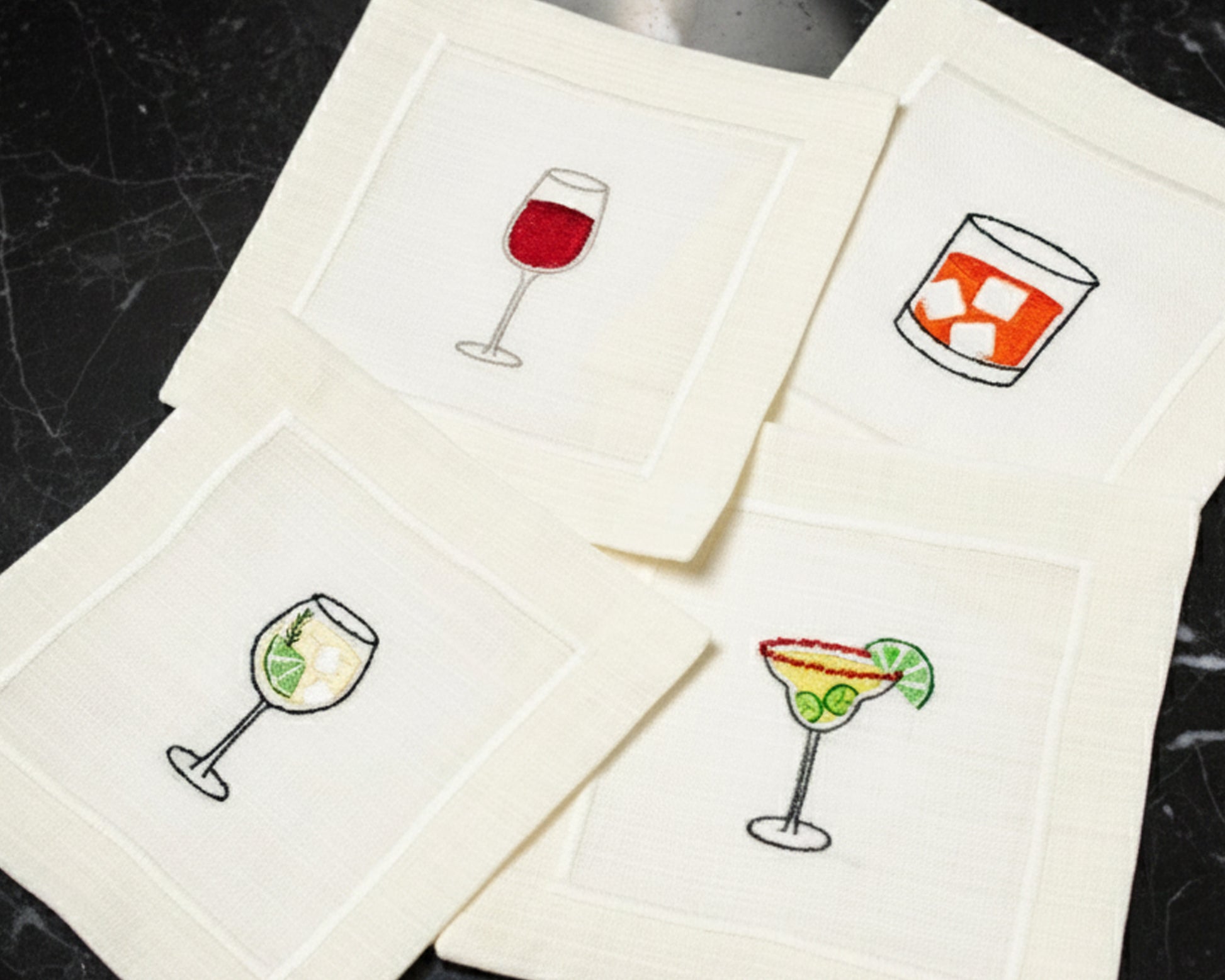 A variety of cream cotton napkins featuring embroidered icons of a Pina Colada, Gin & Tonic, and Aperol Spritz.